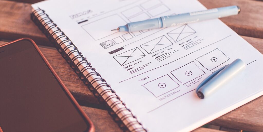 Website wireframe sketch in a notebook next to a smartphone and pen, symbolizing the website planning phase.